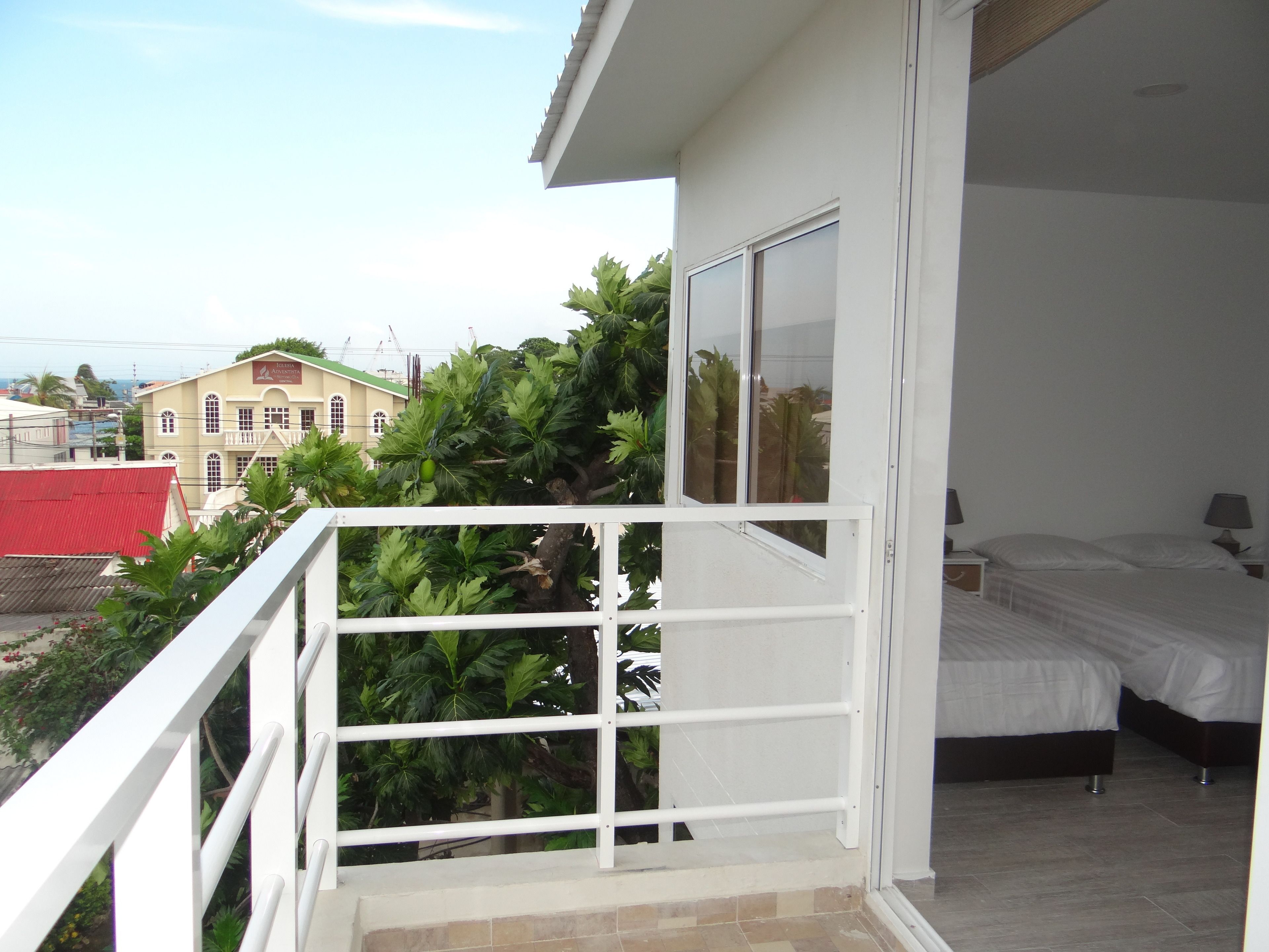 A balcony with a white railing, a bed with white bedding, and a view of a building with a red roof.