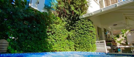 A swimming pool with blue mosaic tiles, a white building with a balcony, and lush greenery.