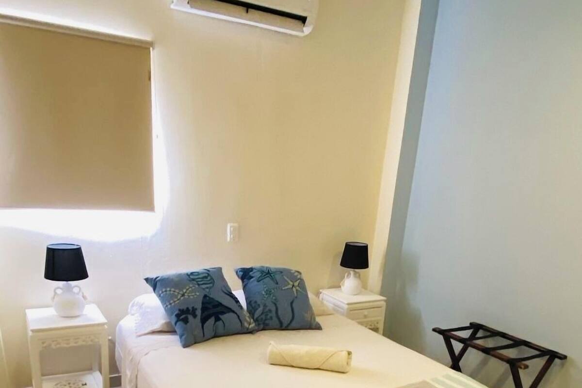 A bedroom with a bed, two bedside tables, a nightstand, an air conditioning unit, a window, and a small table with a lamp.