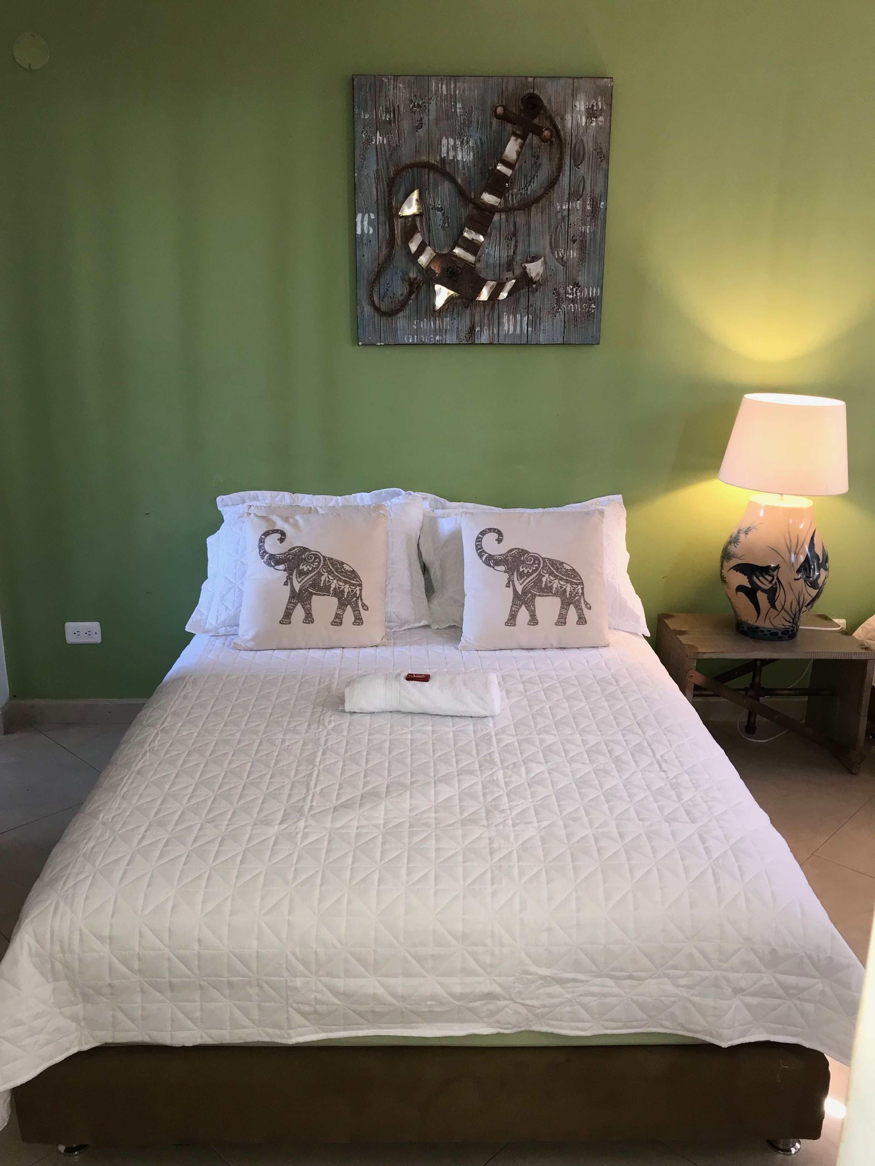A neatly made bed with a quilted white duvet, two pillows with elephant designs, a wooden headboard, a wall-mounted abstract art piece, and a bedside lamp with a floral design.