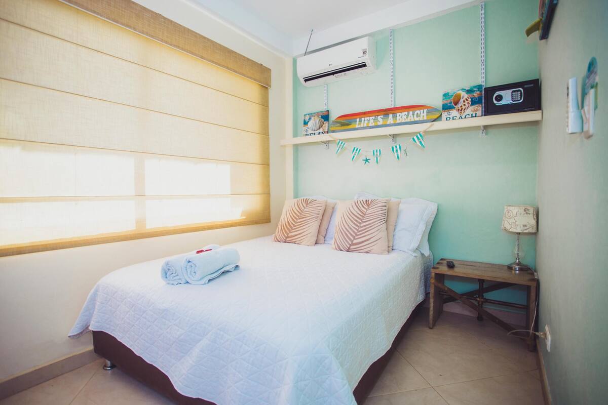 A bedroom with a bed, a window, an air conditioner, a shelf with decorations, and a bedside table.