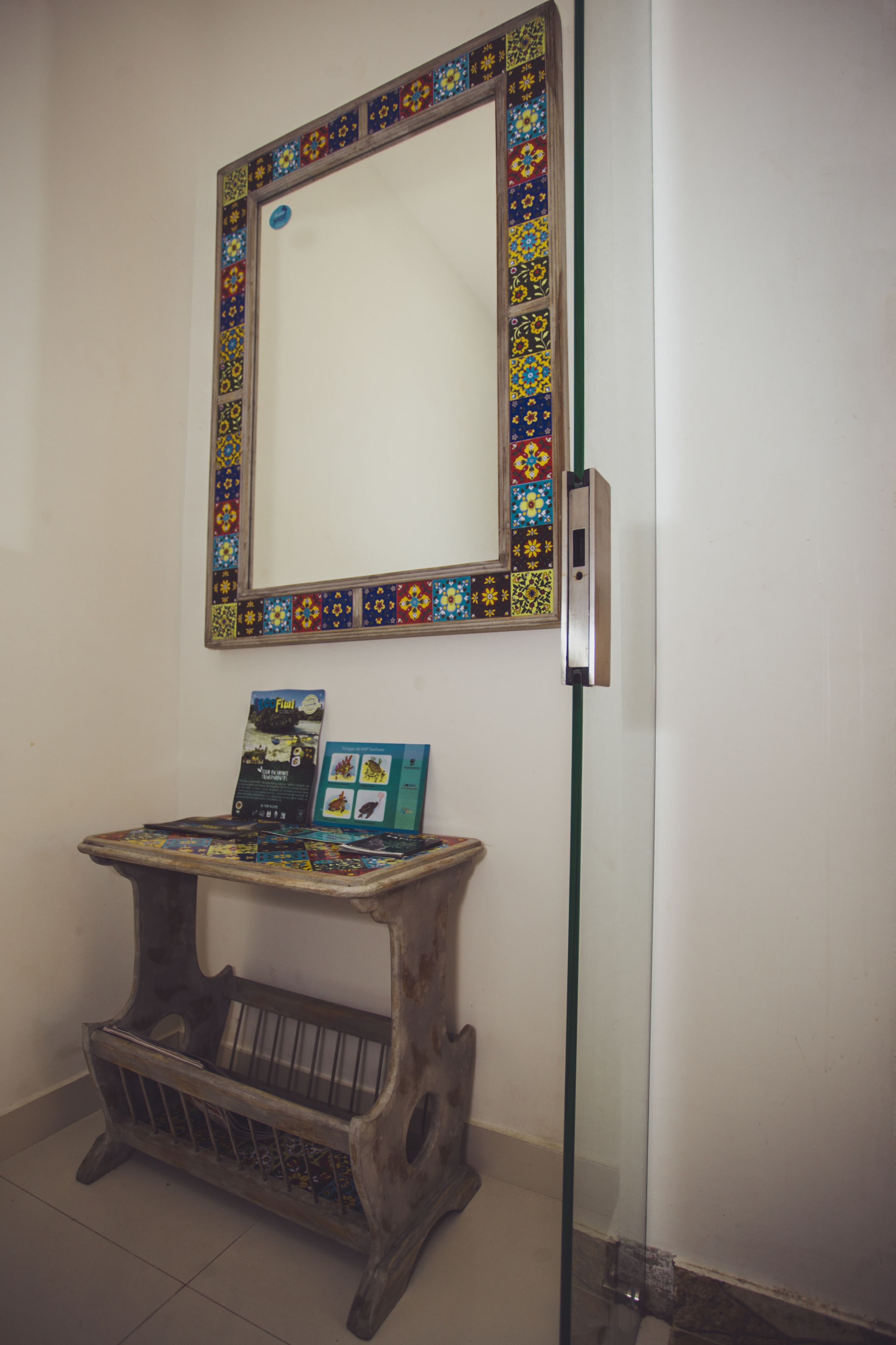 A mirror with a colorful frame, a small table with a book, and a wall-mounted light switch.