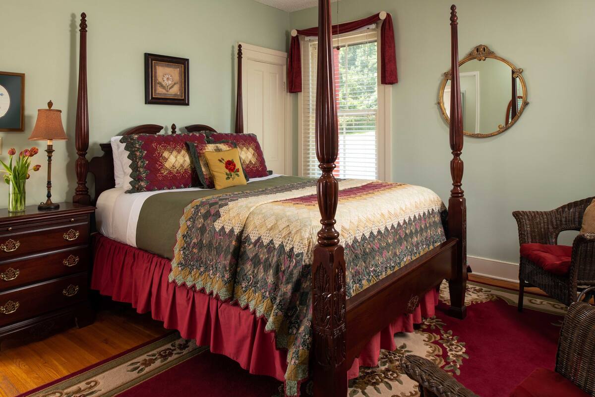 A four-poster bed with a floral bedspread, a wooden dresser with drawers, a wicker chair, and a mirror on the wall.