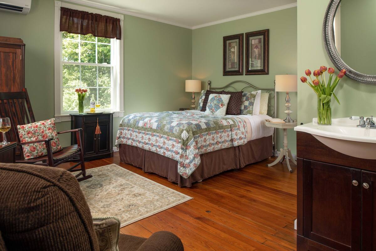 A bedroom with a large bed, bedside tables, a dresser, a window with curtains, and a vase with flowers.