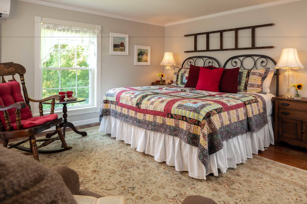 A bedroom with a large bed, a rocking chair, a bedside table with lamps, and a window with curtains.