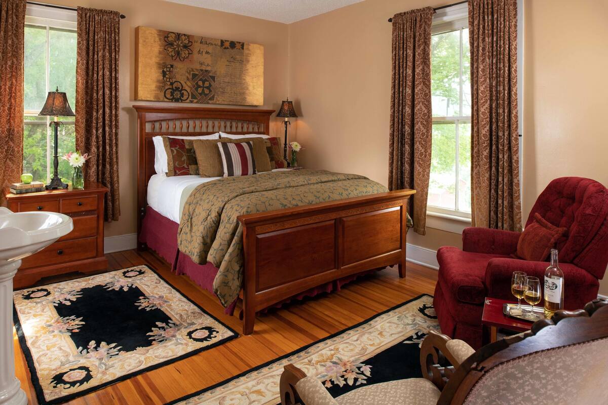 A bedroom with a wooden bed, a red armchair, a nightstand with a lamp, and a window with curtains.