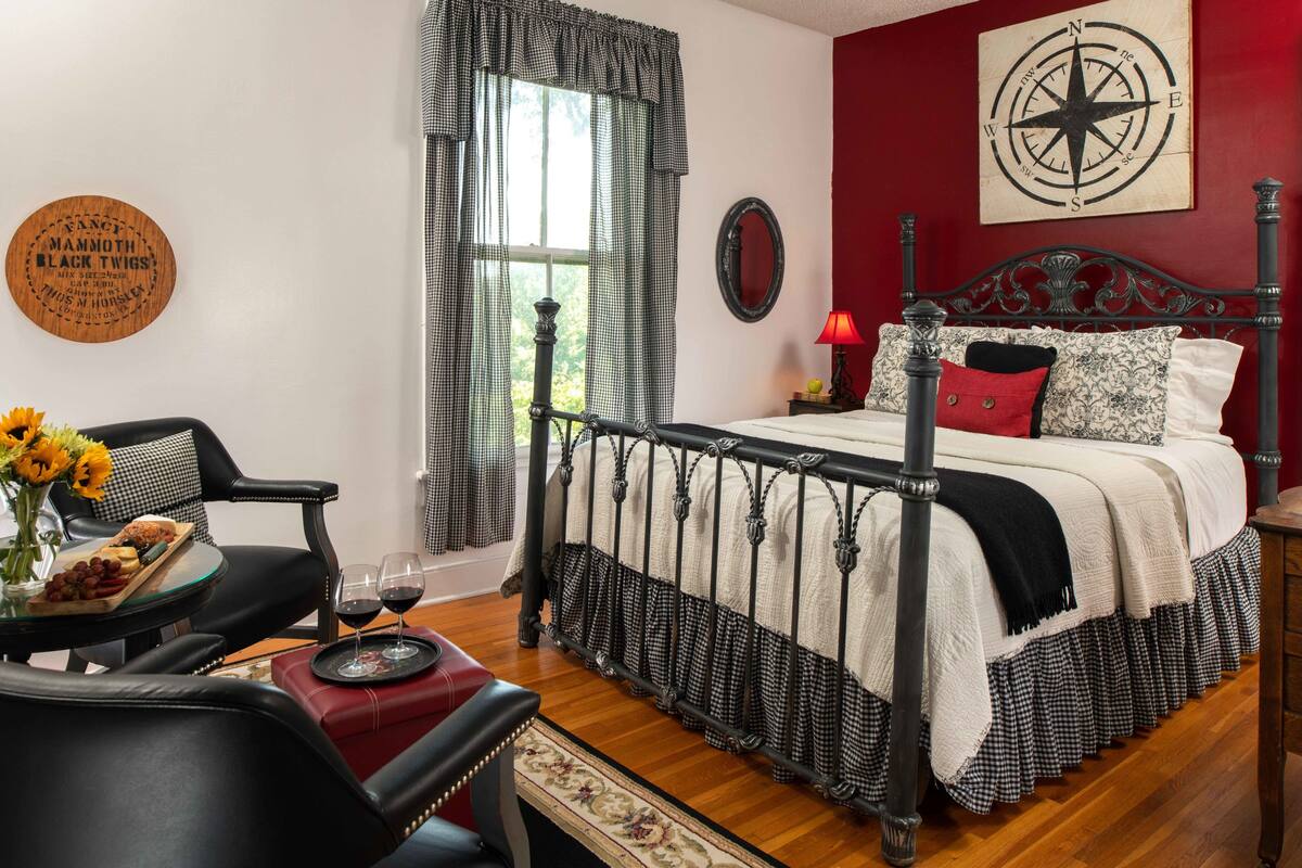A bedroom with a bed, a chair, a table with a tray of food and drinks, a window with curtains, a compass wall decoration, and a round wooden sign.
