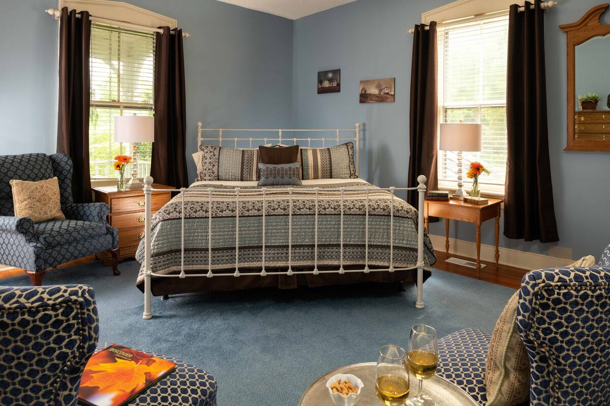 A bedroom with a bed, two chairs, a nightstand, a dresser, and a window with curtains.