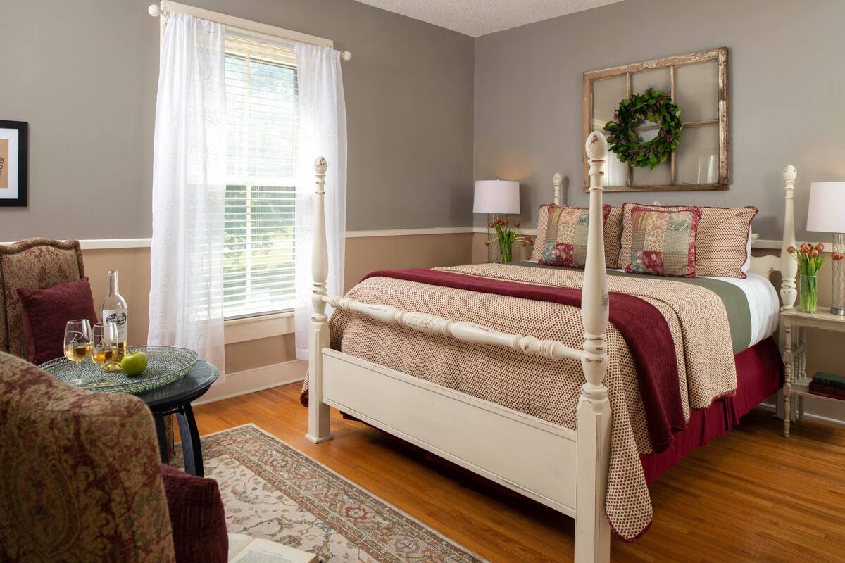 A bedroom with a four-poster bed, a window with sheer curtains, a bedside table with lamps, and a decorative wreath on the wall.