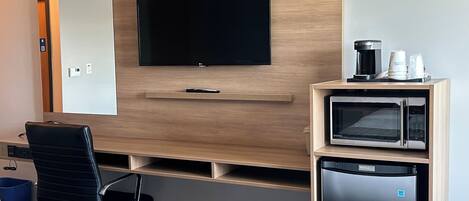 Quarto de hotel com televisão de tela plana fixada em parede de madeira, pequena escrivaninha com micro-ondas e cafeteira, e cadeira de escritório preta.