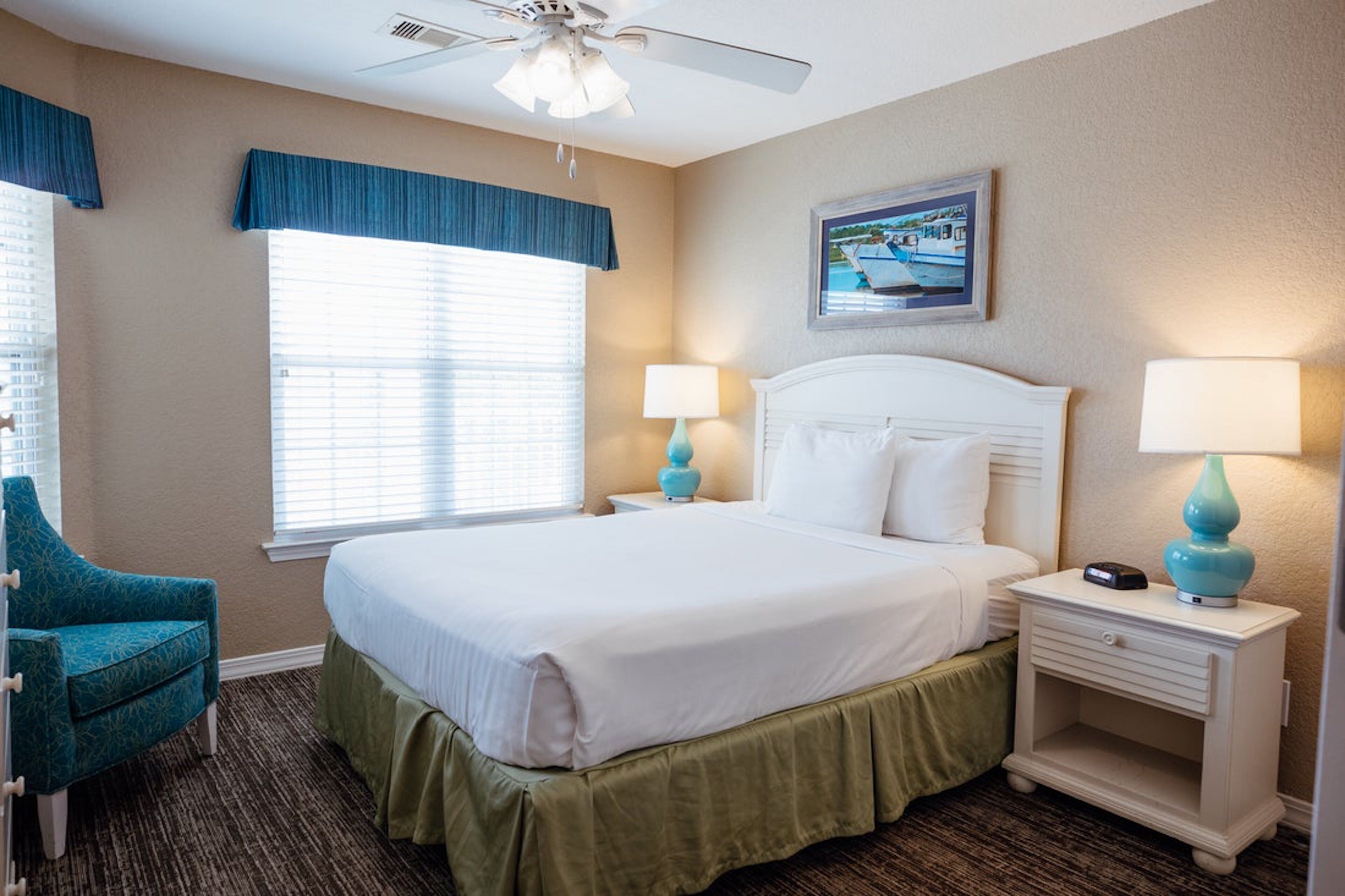 A hotel room with a large bed, two bedside tables with lamps, a ceiling fan, a window with blinds, a chair, and a painting on the wall.