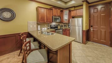 A kitchen with wooden cabinets, a stainless steel refrigerator, a microwave, and a sink. There are two bar stools and a clock on the wall.