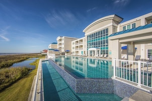 A hotel with a large outdoor swimming pool, a covered area with lounge chairs, and a view of the ocean.