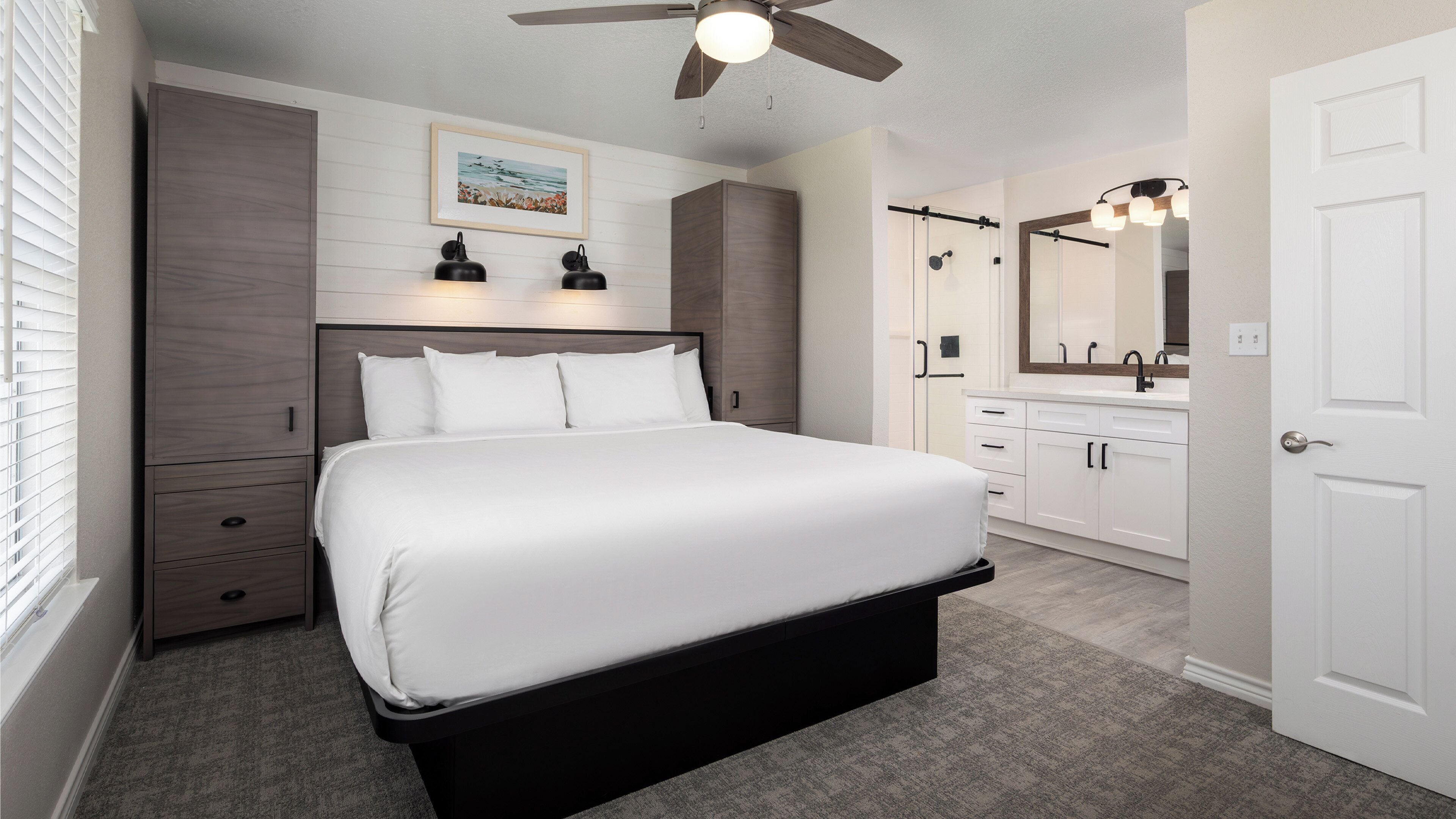 A bedroom with a large bed, a ceiling fan, a bathroom with a mirror and sink, and a window with blinds.