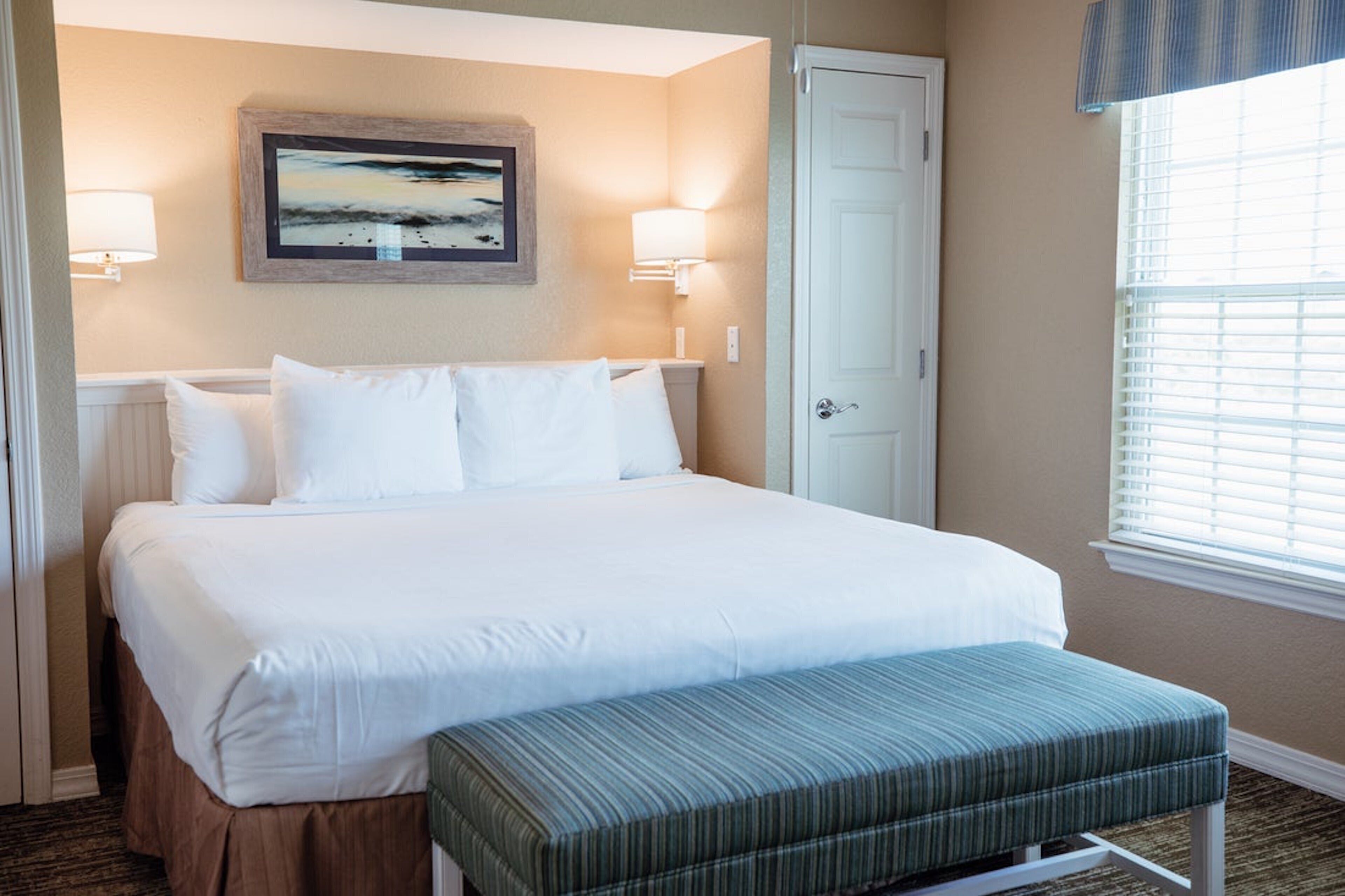 Habitación de hotel con una cama grande, un banco, dos lámparas de pared, una puerta y una ventana con persianas.