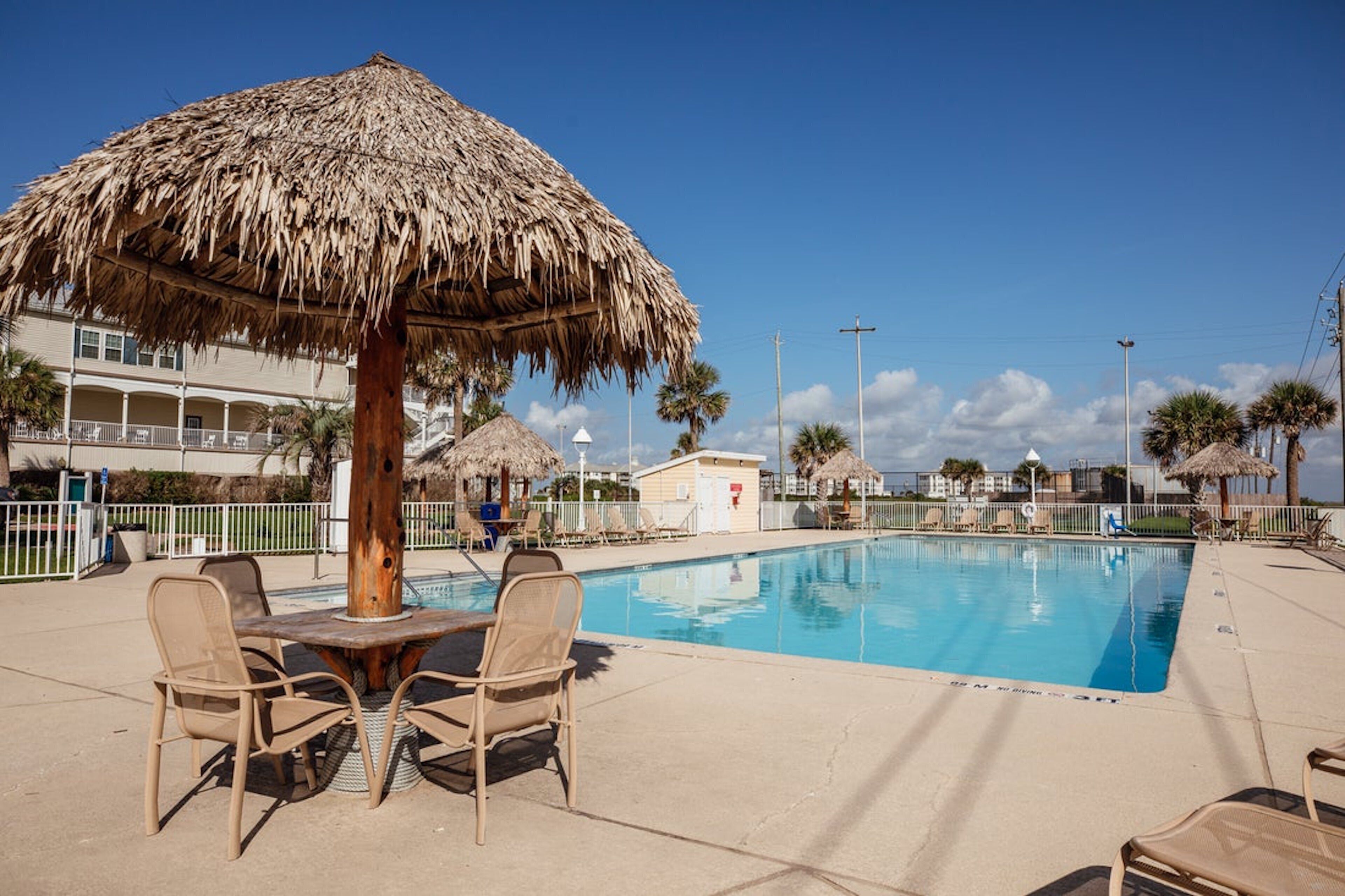 A pool area with a thatched umbrella, lounge chairs, and a building in the background.