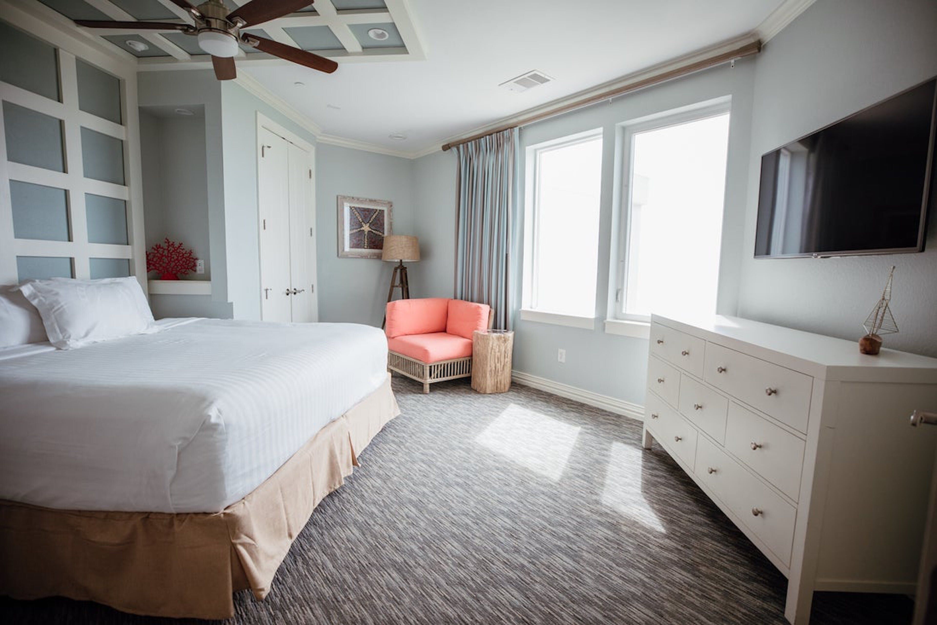 Une chambre d’hôtel avec un grand lit, une chaise, une commode, une télévision et une fenêtre avec des rideaux.