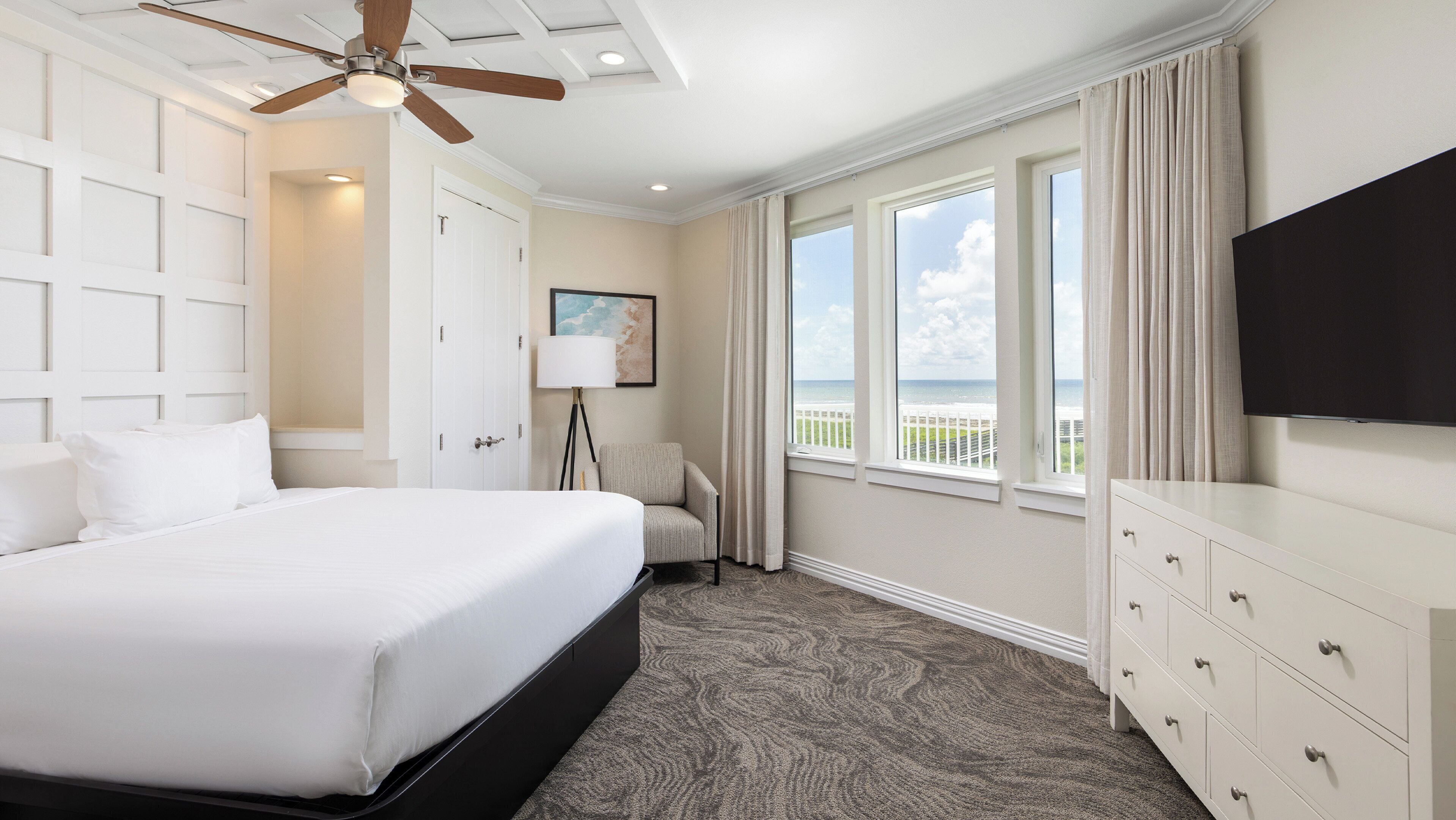 A hotel room with a large bed, a ceiling fan, a desk with a TV, and a view of the beach.