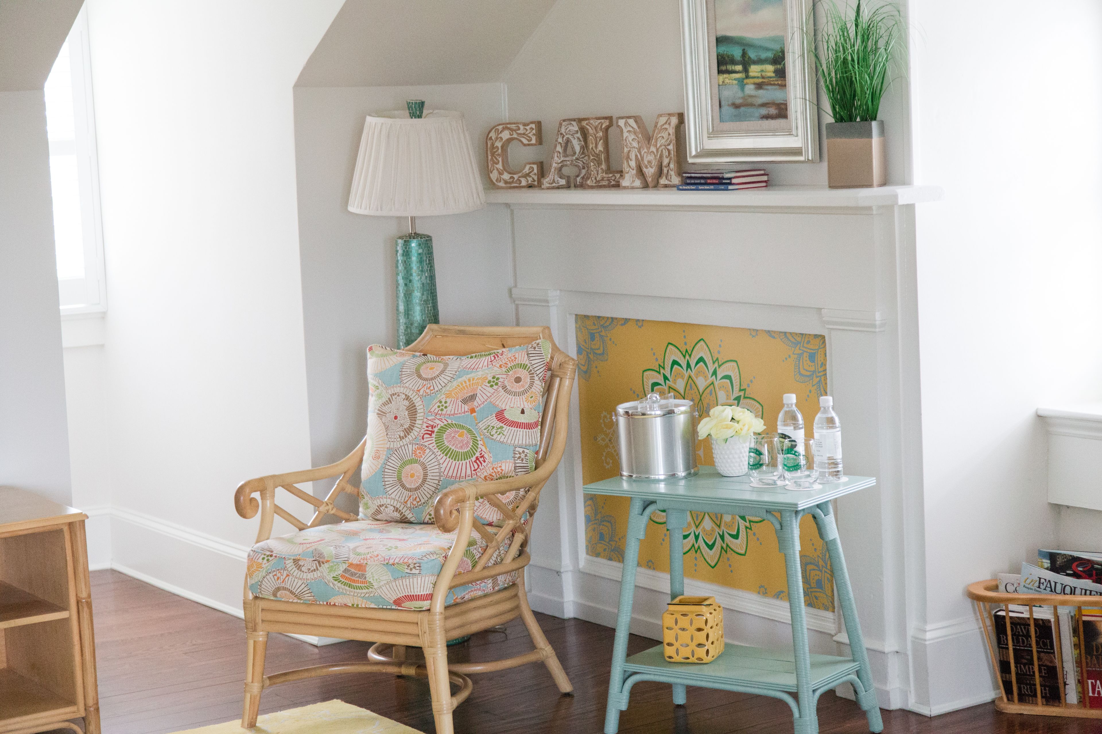 A cozy room with a wicker chair, a small table with decorative items, and a fireplace with a 'CALM' sign above it.