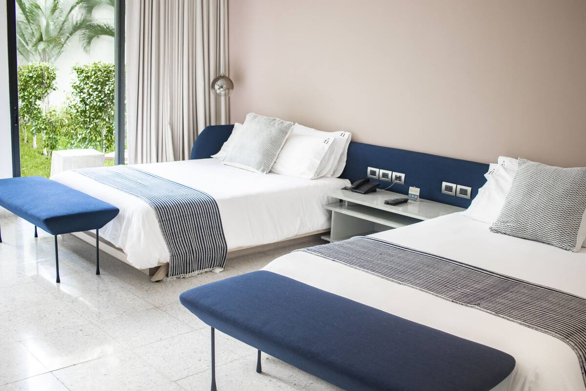 A hotel room with two beds, a bench, a bedside table, and a view of greenery outside.