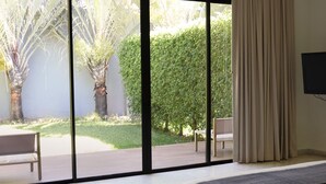 A bedroom with a large sliding glass door leading to a patio area with a bench, a small table, and a view of a neatly trimmed hedge and palm tree. - Il Buongustaio (San Salvador)