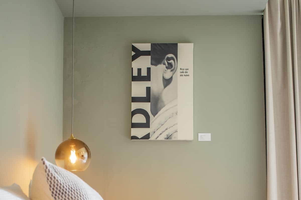 A neatly made bed with white bedding and decorative pillows, a hanging pendant light, and a framed artwork on the wall.