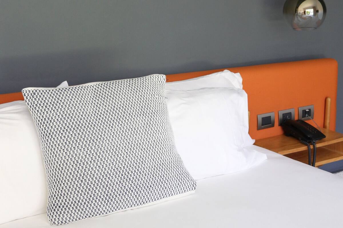A hotel room with a bed, a patterned pillow, a white pillow, and a bedside table with a phone.
