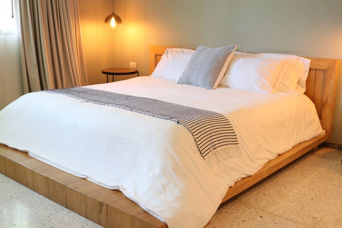 A neatly made bed with a white duvet and a patterned throw, a small bedside table, and a wall-mounted light fixture.