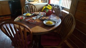 A wooden dining table with a placemat, surrounded by wooden chairs. There is a bowl of fruit, plates with food, and a vase with flowers on the table. - Southern Bluff B&B (Soldotna)