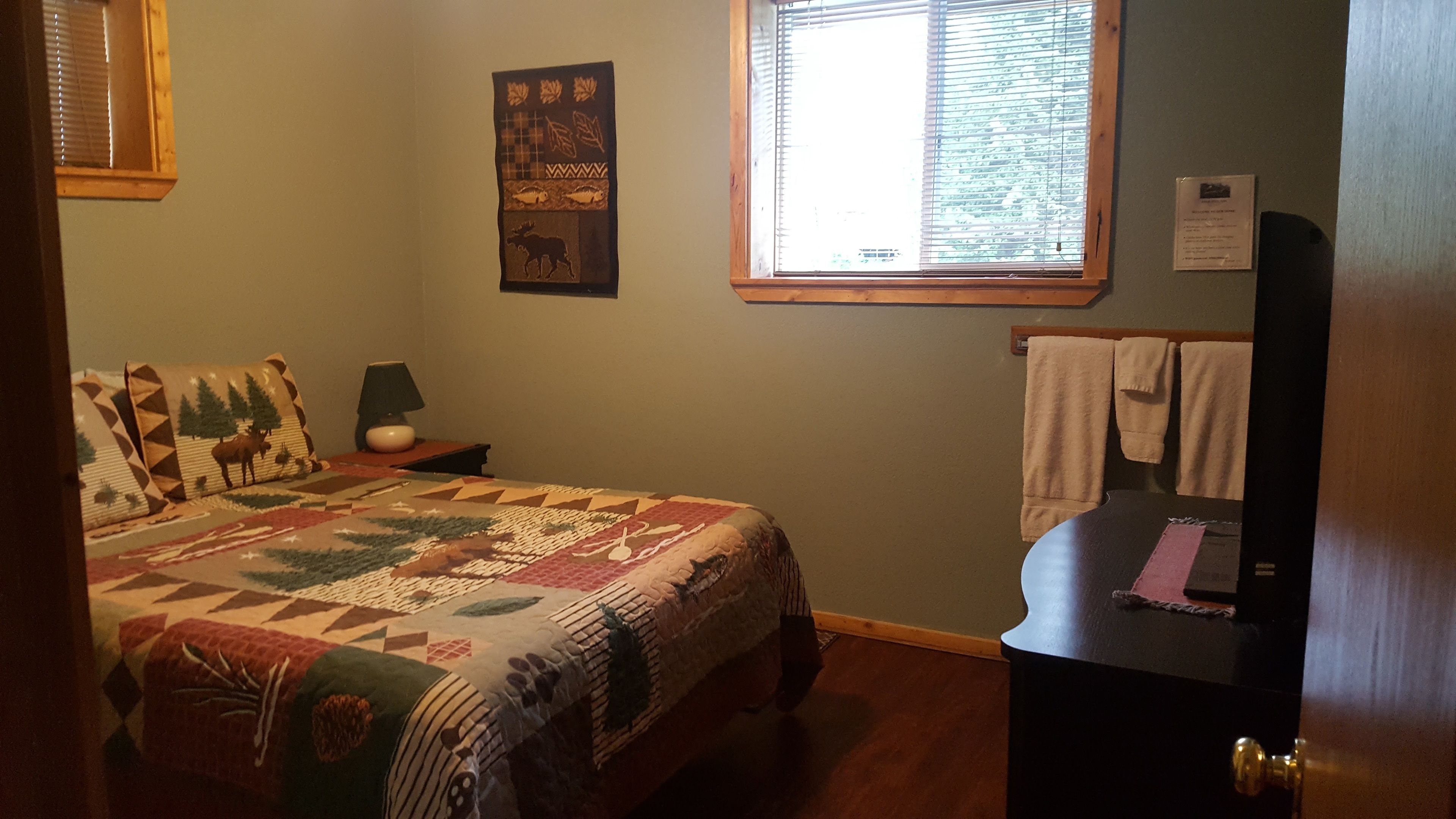 A bedroom with a bed, a nightstand, a lamp, a window with blinds, a towel rack, and a door.