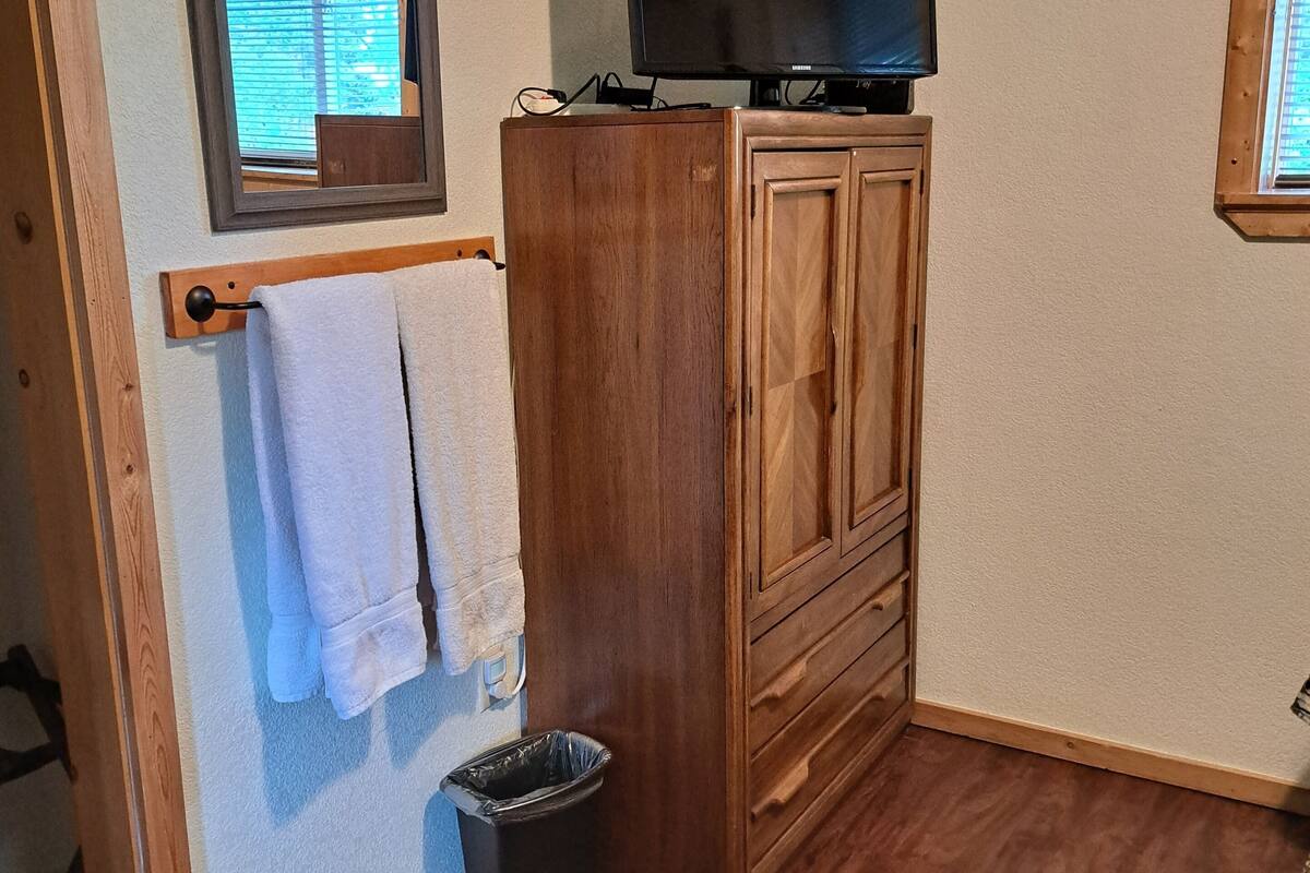 A room with a wooden cabinet, a television on top, a window with blinds, a towel hanging on a rack, and a trash bin.