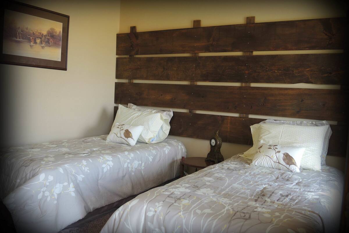 A room with two beds, each with floral-patterned bedding and matching pillows. There is a wooden headboard above the beds, and a clock on the nightstand beside one bed.