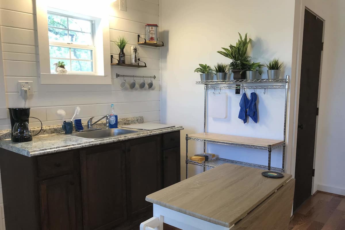 A kitchen with dark wooden cabinets, a granite countertop, a coffee maker, a sink, and a small table. There are potted plants on a shelf above the sink and a towel rack with a blue towel.