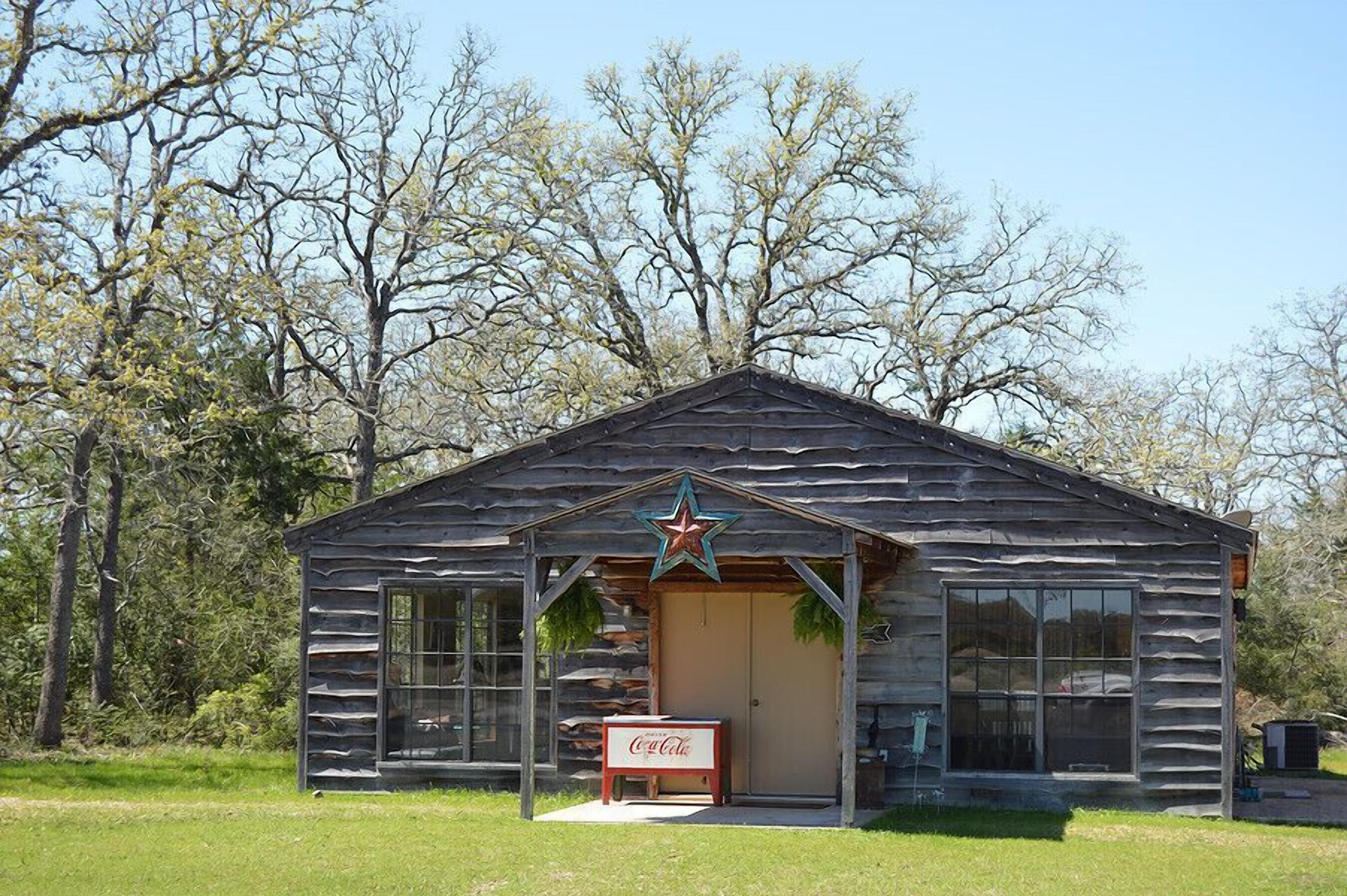 A rustic log cabin with a star decoration on the roof, a Coca-Cola sign in front, and a large window on the side.