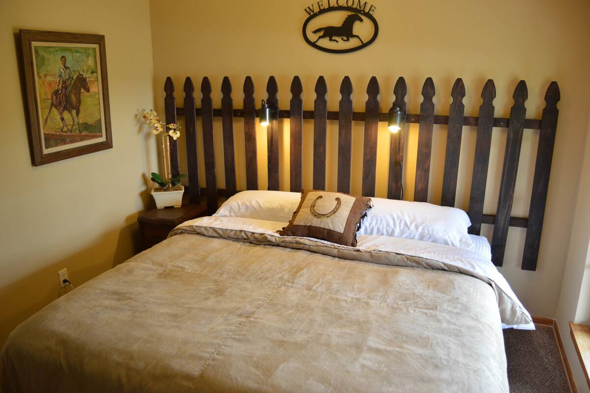 A bed with a wooden headboard, a framed painting, a potted plant, and a 'WELCOME' sign.