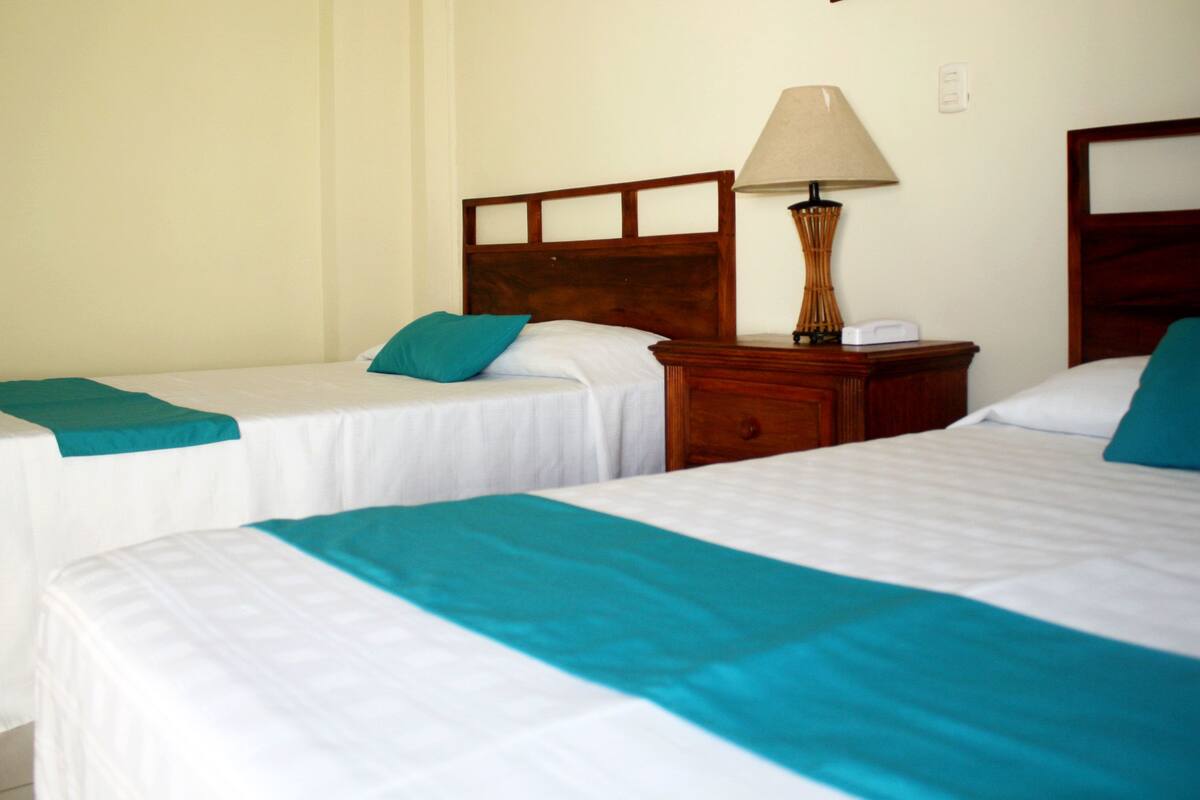 A hotel room with two single beds, each with a teal bedspread and white linens. There is a wooden nightstand between the beds with a lamp and a small white device.