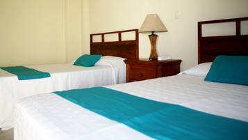 A hotel room with two single beds, each with a teal bedspread and white linens. There is a wooden nightstand between the beds with a lamp and a small white device.