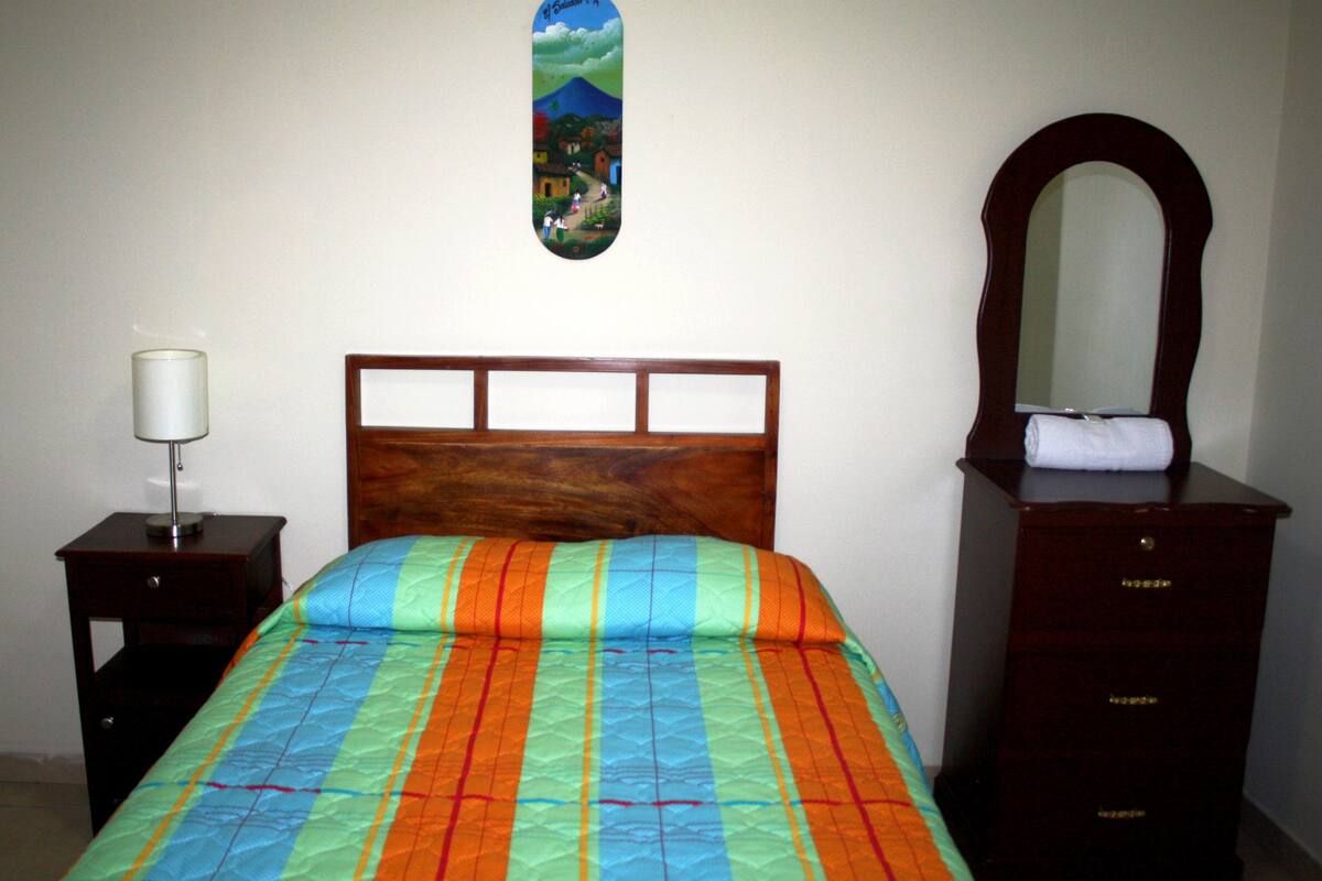 A bedroom with a wooden headboard, a colorful bedspread, a bedside lamp, a dresser with a mirror, and a wall-mounted skateboard.