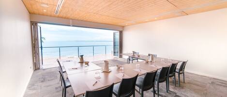 A conference room with a large table and chairs, a view of the sea through a glass door, and a wooden ceiling.