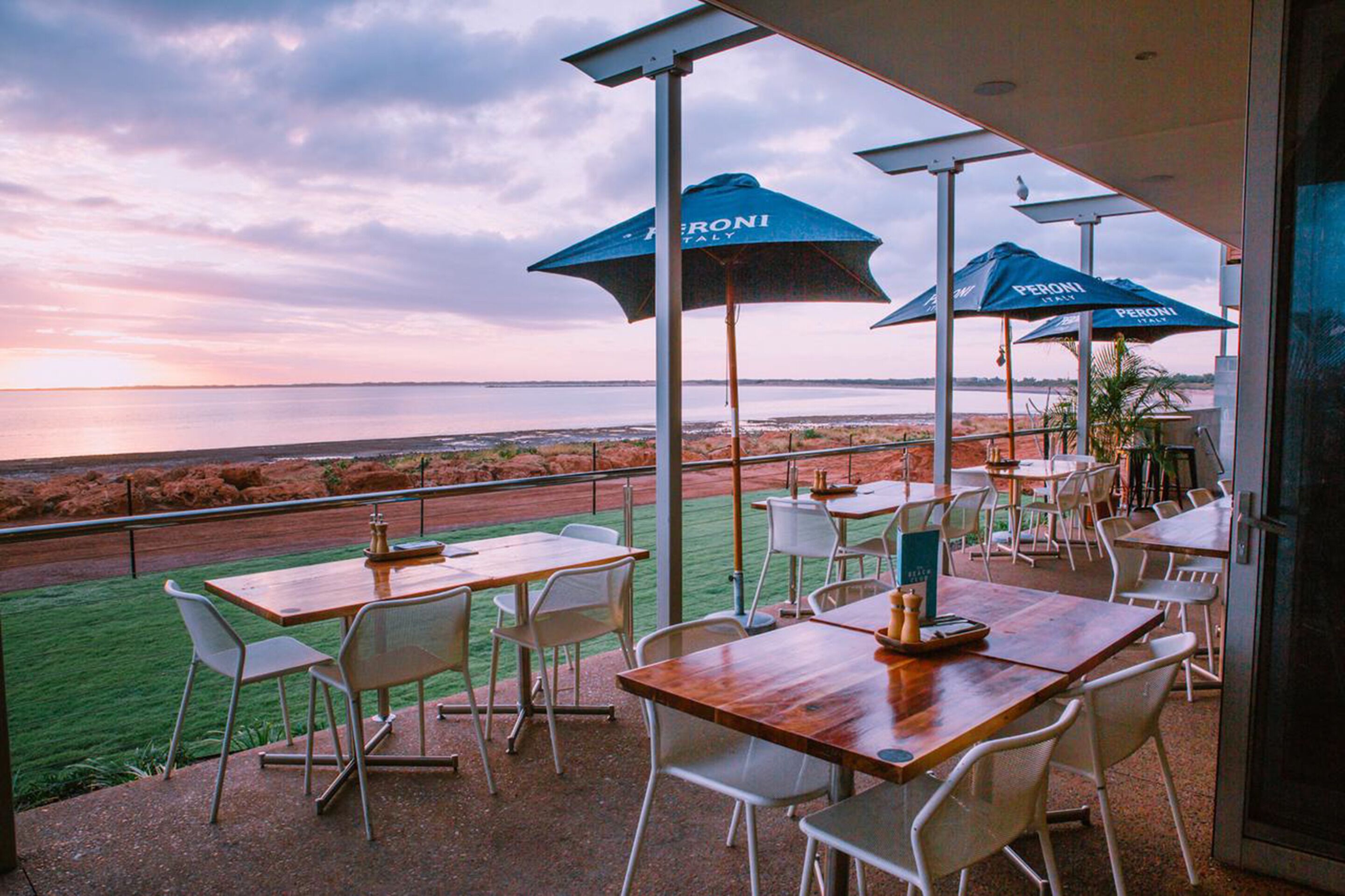 An outdoor dining area with tables and chairs, blue umbrellas, and a view of the sea.