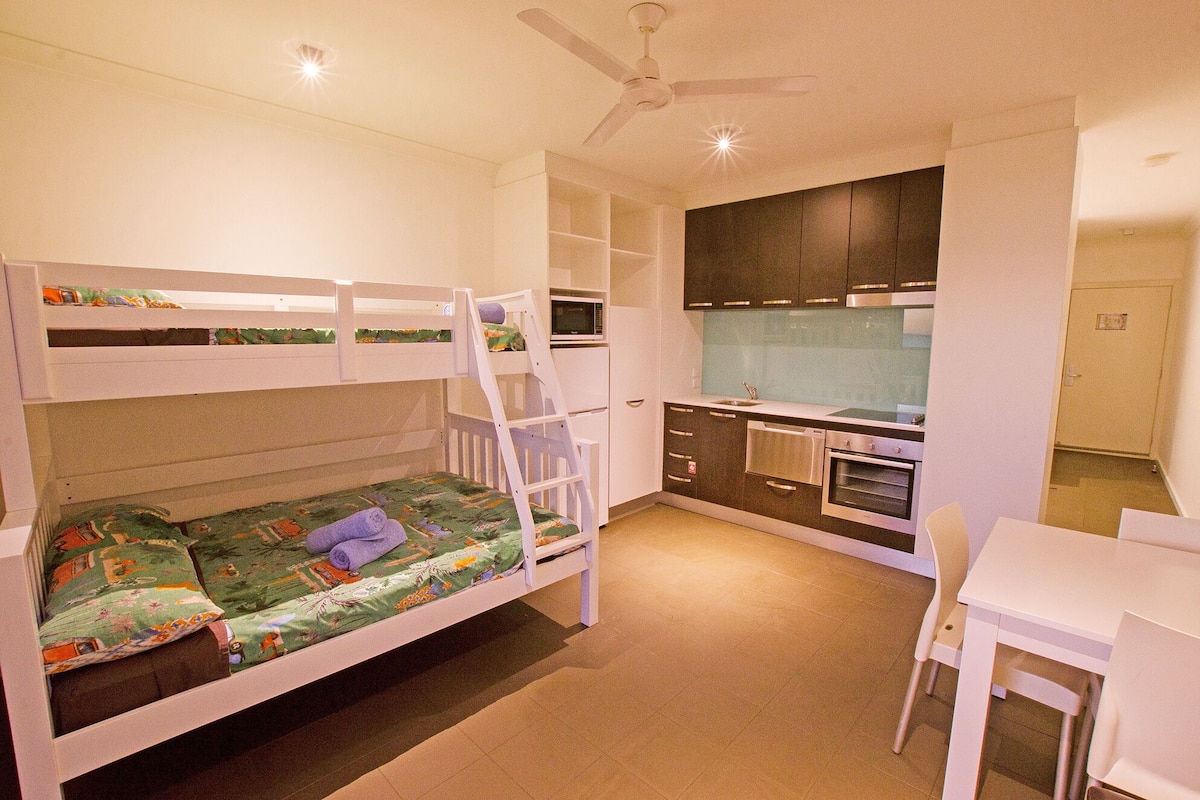 Uma cama de beliche com escada, uma cozinha compacta com micro-ondas e uma mesa branca com cadeira.