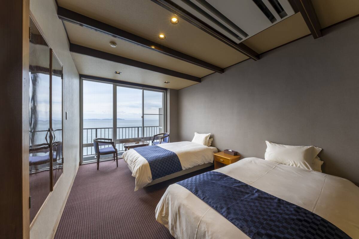 A hotel room with two beds, a small table, a chair, and a balcony with a view of the sea.