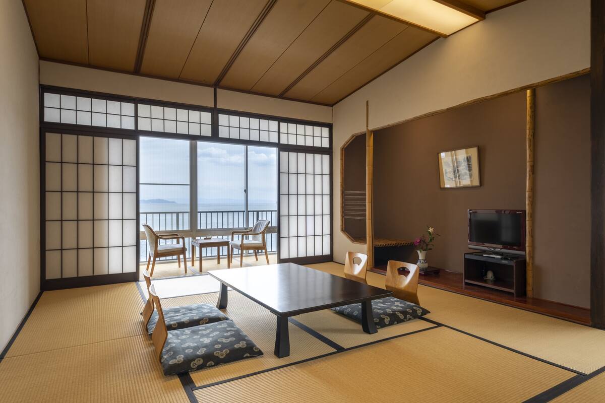 A traditional Japanese room with tatami flooring, a low table, chairs, a television, and a view of the sea.