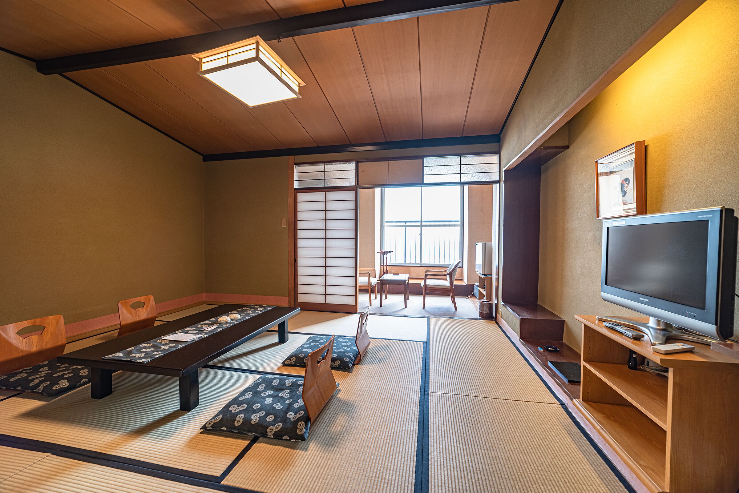 Et traditionelt japansk rum med tatamigulv, et lavt bord, træstole, et fjernsyn og skydedøre, der fører til en balkon.