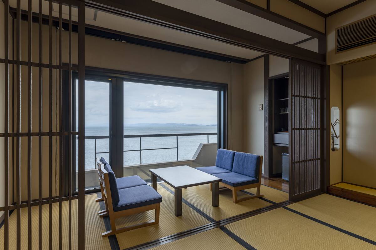A traditional Japanese room with tatami flooring, a low table, two blue cushioned seating areas, and a view of the sea through large windows.