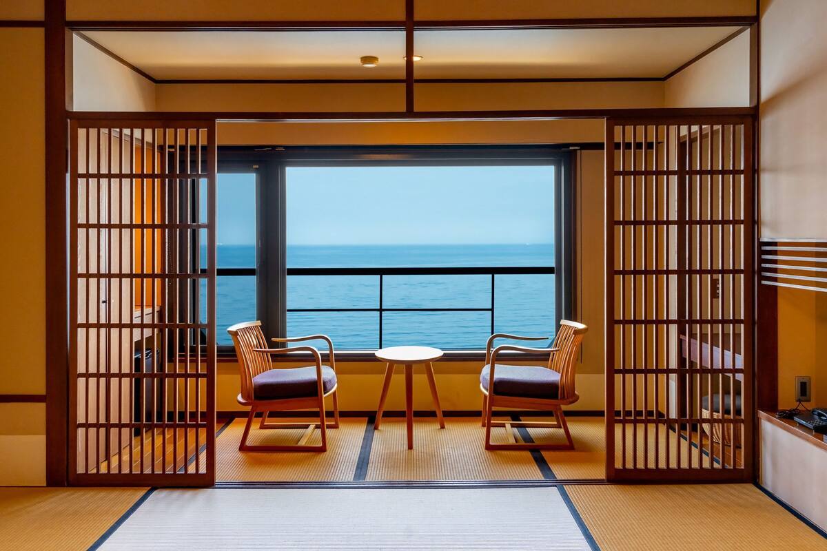 A room with tatami flooring, a low table with two chairs, and a view of the sea through large windows.