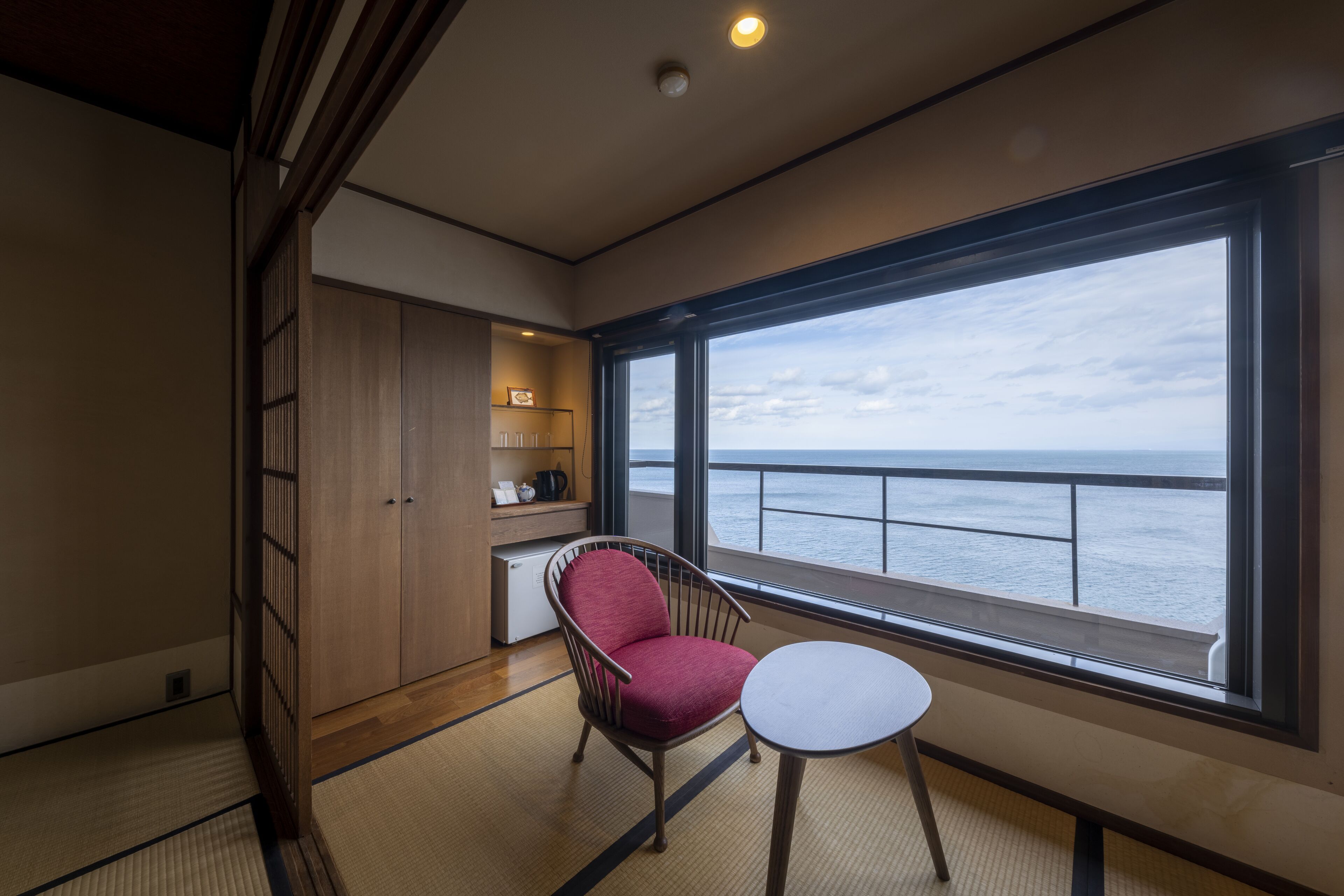A room with a view of the sea, a red chair, a small table, and a cabinet with shelves.