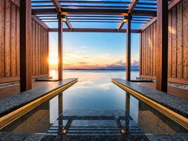 Zona de spa con paredes de madera y piso de vidrio que da a una piscina, reflejando el atardecer.