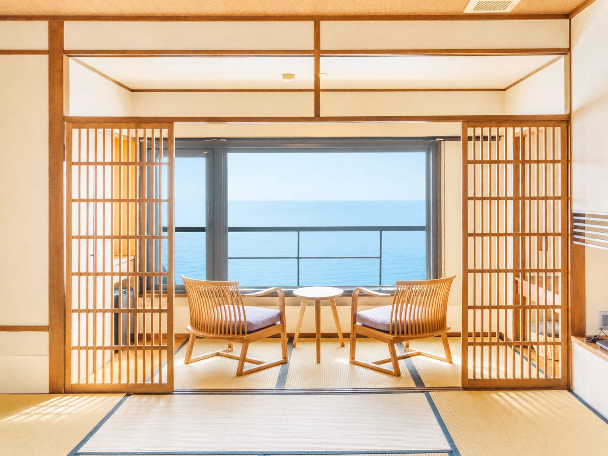 Une pièce de style japonais traditionnel, avec un sol recouvert de tatamis, une petite table basse, deux chaises et une vue sur la mer par une grande fenêtre.