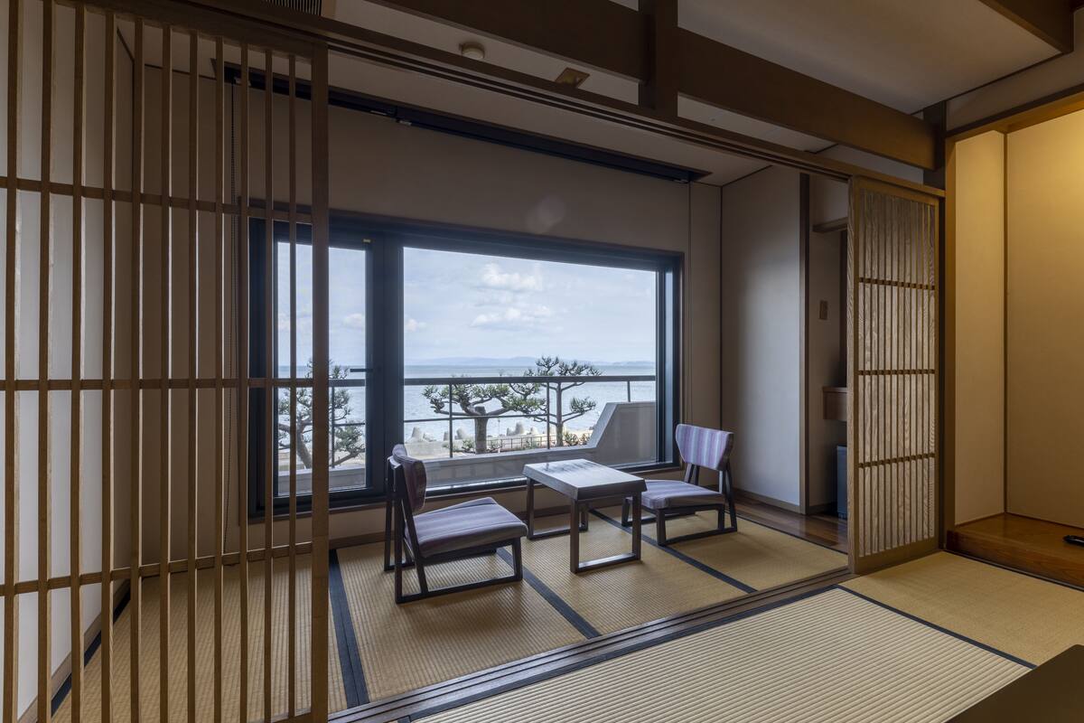 A traditional Japanese room with tatami flooring, a low table, two chairs, and a view of the sea.